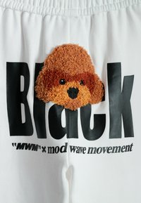 Pantalones de chándal blancos con un estampado gráfico negro que dice "Black" y un parche de cabeza de perro marrón y peludo situado encima del texto.