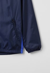 Navy windbreaker jacket with a smooth texture, features a side zip pocket, elastic cuffs, and a blue perforated accent on the side.