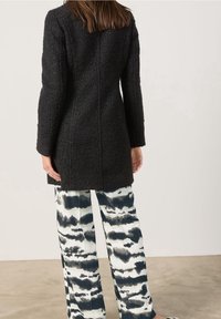Sort tekstureret frakke med lange ærmer og en struktureret form, båret over løstsiddende, mønstrede bukser i hvide og mørkeblå nuancer.