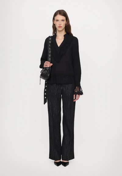 Bluză neagră cu guler volanat; pantaloni negri cu dungi, cu o croială lejeră; ține în mână o poșetă mică, neagră; tocuri negre elegante.