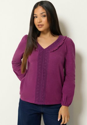 Mujer con cabello largo y oscuro, que lleva una blusa morada de manga larga con detalles de encaje y pantalones azul oscuro sobre un fondo claro y liso.