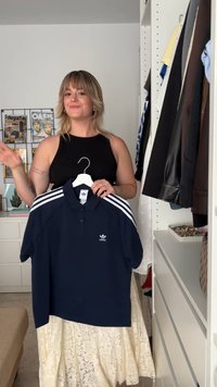 Camisa polo azul-escuro com três riscas brancas nos ombros e logótipo, feita de tecido com textura suave, pendurada num cabide branco.