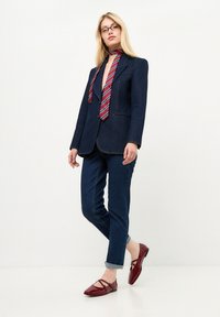 Marineblaues Denim-Blazer mit Kontrastnähten, kombiniert mit dunklen Jeans und glänzenden burgunderroten Ballerinas. Eine gestreifte rot-blaue Krawatte rundet den Look ab.