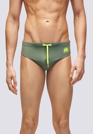 Torace e gambe maschili che indossano slip da nuoto verde oliva con cordoncino giallo neon e logo sul lato destro, in piedi su sfondo grigio chiaro.