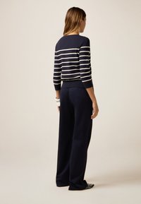Donna in piedi di spalle, vestita con un maglione a righe blu navy e bianco, pantaloni larghi blu navy e scarpe a punta con borchie.