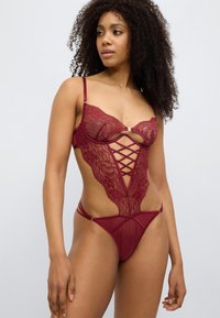 Κορμάκι από δαντέλα σε μπορντό με ρυθμιζόμενες τιράντες, που διαθέ τει περίπλοκες κοψίματα και σχέδιο σε σχήμα V, αναδεικνύοντας υφή και λεπτομέρειες.