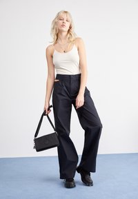 Sorte vidde bukser, beige tanktop, sort håndtaske med strop. Glat stof, minimalistisk design. Model står foran en blå baggrund.