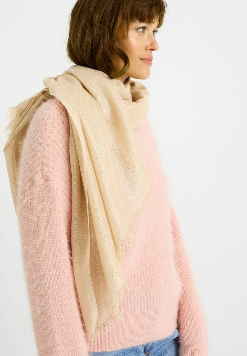 Écharpe beige avec un léger scintillement, présentant une texture douce et des bords à franges, drapée élégamment sur un pull rose clair texturé.
