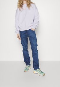 Ljuslila sweatshirt med ett broderat detalj, blå jeans och färgglada sneakers med en texturerad mesh-överdela och gummisula.