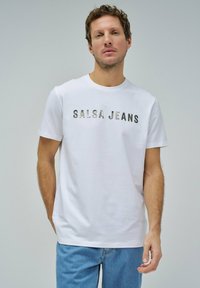 Camiseta de algodón blanca con la inscripción "SALSA JEANS" en letras metálicas, diseño de cuello redondo, mangas cortas y un ajuste relajado, combinada con jeans de un azul claro.