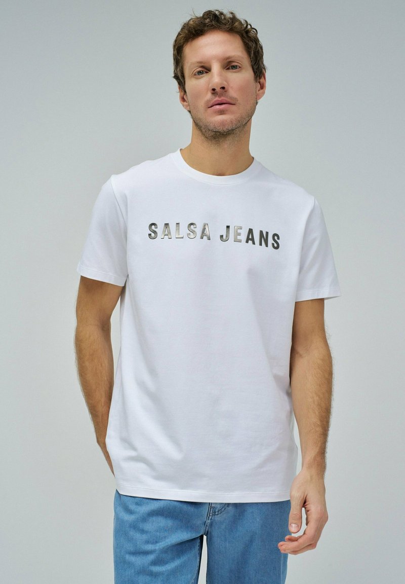 Camiseta de algodón blanca con la inscripción "SALSA JEANS" en letras metálicas, diseño de cuello redondo, mangas cortas y un ajuste relajado, combinada con jeans de un azul claro.