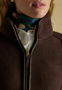 Kobieta ubrana w brązową kurtkę na zamek, pod którą ma białą koszulę oraz zielony kwiatowy szal na szyi, usta częściowo widoczne.