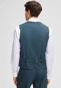 Mężczyzna widziany z tyłu, ubrany w dopasowaną, ciemnozieloną kamizelkę i pasujące spodnie, pod którymi ma białą koszulę z długim rękawem.