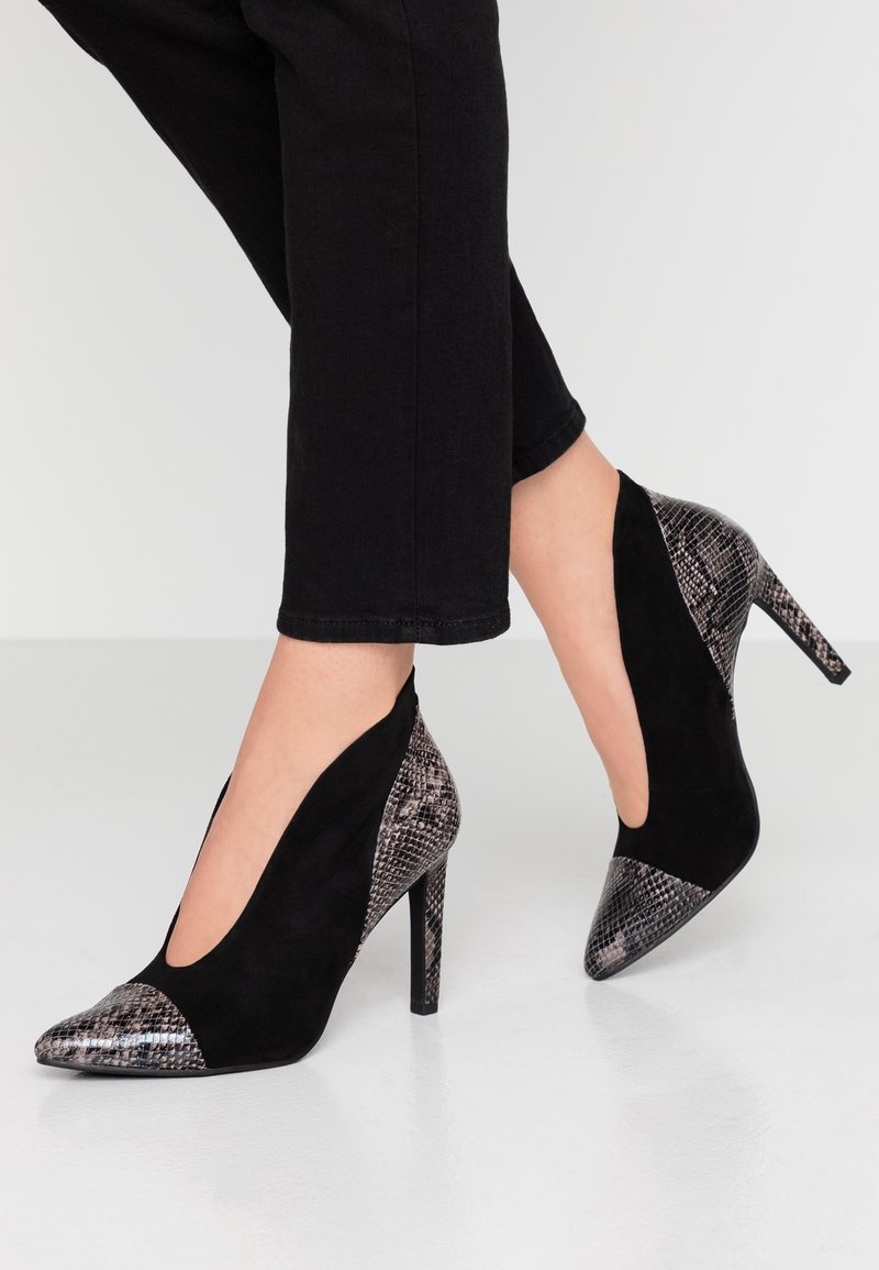 Chaussures à talons hauts noires avec bouts pointus et accents imprimé serpent sur les talons, les bouts et l'arrière, portées avec un pantalon court noir.