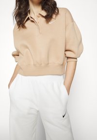 Beige Sweatshirt mit einem Polokragen, drei Knöpfen und einem weißen Nike-Logo auf der Brust, kombiniert mit weißen Sweatpants, die ein kleines Logo aufweisen.