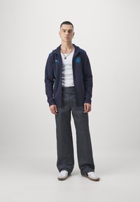 Námornícky mikina s tyrkysovými akcentmi, zips vpredu, logo na hrudi; biela tričko bez rukávov; šedé široké nohavice; biele tenisky s hnedými podrážkami.