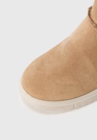 Pointe de bottes en daim beige avec semelle en caoutchouc blanc sur fond blanc uni, montrant la texture et la couture détaillées.