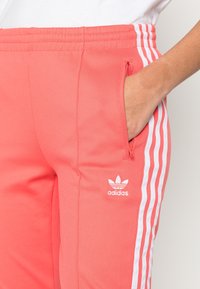 Pantaloni da atletica color corallo con strisce laterali bianche e logo. Presentano una cintura elastica, una tasca con zip e una superficie liscia.