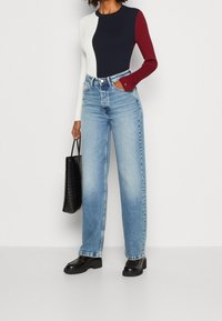 Donna che indossa una maglietta a maniche lunghe color block bianca, blu navy e bordeaux, jeans a vita alta azzurri chiari, scarpe nere e tiene una borsa tote nera.