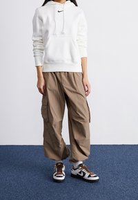 Persona vistiendo una sudadera blanca de Nike, pantalones cargo marrones sueltos y zapatillas Nike negras, blancas y marrones, de pie sobre una alfombra azul.