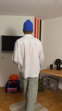 Bílá pruhovaná košile, volného střihu s švem na zádech; v kombinaci s světle zelenými kalhotami. Doplněno modrou pletenou čepicí, stojící v místnosti.