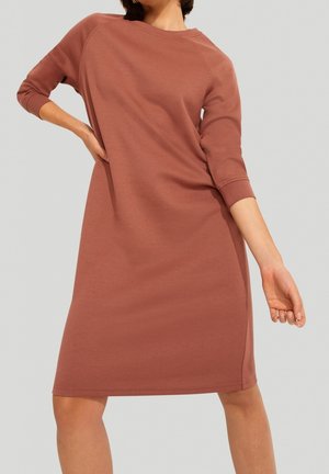 Vrouw in een knielange, stofroze jurk met lange mouwen, poserend met één hand aan haar heup en de andere ontspannen langs haar zij.