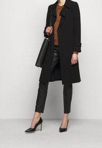 Schwarzer Mantel mit langem Design und leicht umgeschlagenem Kragen, kombiniert mit braunem Pullover, schwarzen Lederhosen und spitzen schwarzen High Heels.