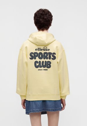 Personne aux cheveux courts et bouclés portant un sweat à capuche jaune pâle avec l'inscription « ellesse SPORTS CLUB since 1959 » et une jupe en jean bleue, vue de dos.