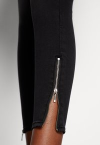 Leggings de denim pretos apresentam zíperes nos tornozelos, acessórios prateados e um design justo. O tecido parece suave com um ligeiro estiramento.