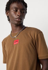 Ung mann iført en brun t-skjorte med en rød "HUGO" logo-patch og et sølv kjedehalskjede, ser ned og til siden.