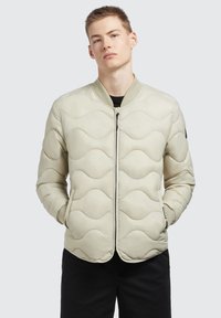 Beige quiltad bomberjacka med vågmönster, dragkedjestängning, ribbade ärmslut och fickor. Slätt, lättviktigt tyg.