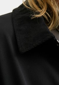 Giacca nera con colletto in corduroy testurizzato e corpo in tessuto liscio, che mette in evidenza un forte contrasto nei materiali e nelle finiture.
