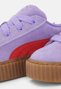 Fialové semišové tenisky s červeným akcentem, kulatou špičkou, texturovanou hnědou gumovou podrážkou a sladěnými fialovými tkaničkami, s vyvýšeným logem na jazyku.