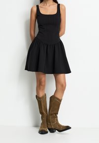 Robe noire sans manches avec un corsage ajusté et une jupe évasée. Associée à des bottes de cowboy en daim marron montant jusqu'au genou, ornées de clous argentés.