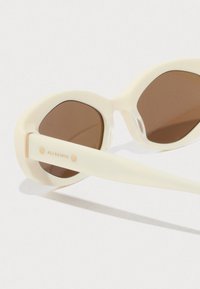 Óculos de sol redondos de cor creme com lentes castanhas e a marca "ALI SAINTS" na haste, colocados sobre uma superfície branca.