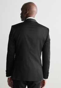Chaqueta de traje negra con corte recto, fabricada en un tejido brillante y con detalles en los botones. Presenta un cuello de camisa blanca visible en el cuello.