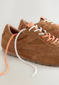 Braune Wildleder-Sneaker mit niedrigem Profil, ausgestattet mit orangefarbenen und weißen Schnürsenkeln, strukturierten Gummisohlen und einem Lederakzent auf der Zunge.