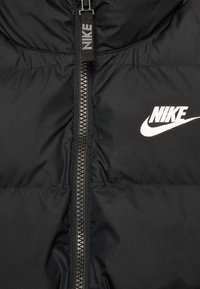 Černá péřovka s předním zipem, která má výrazné bílé logo Nike na levé straně. Hladký, matný povrch.