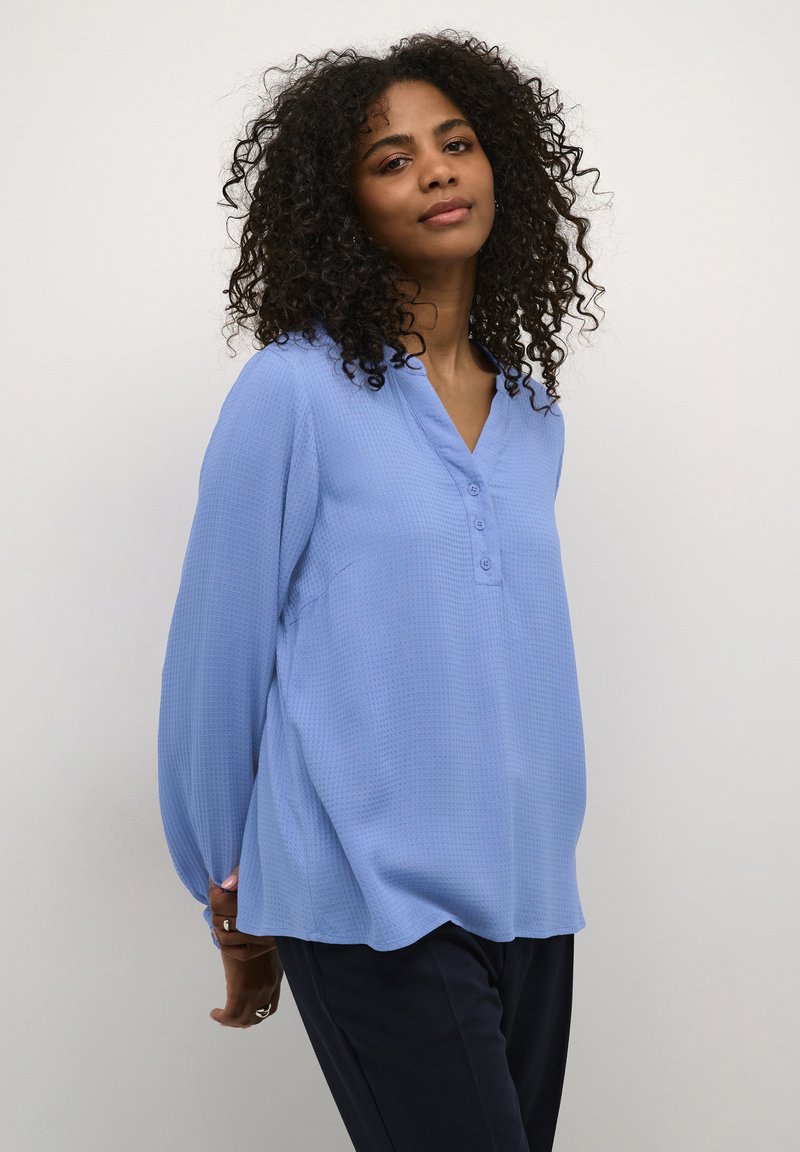 Blouse bleu clair à manches longues avec un décolleté en V et une patte de boutonnage à trois boutons. Tissu texturé avec une coupe décontractée, assortie à un pantalon sombre.