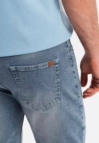 Jeans de mezclilla azules claros con un bolsillo trasero con una etiqueta de cuero marrón y detalles de costura visibles. Textura suave, diseño ajustado.