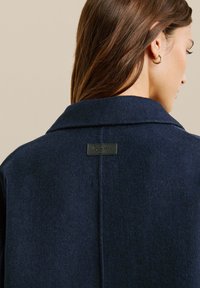 Marineblauwe wollen jas met een geknipt kraag, zichtbare stiksels en een leren merklabel op de achterkant. Gladde textuur.