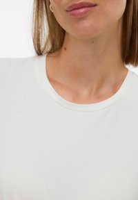 Primer plano del cuello y la mandíbula de una mujer que lleva una camiseta de cuello redondo blanca, con el cabello castaño claro visible a un lado.