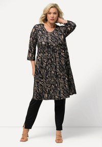 Zwarte jurk met beige bladerenpatroon, knie-lang, en driekwart mouwen, gecombineerd met zwarte broeken en sandaaltjes in tan.