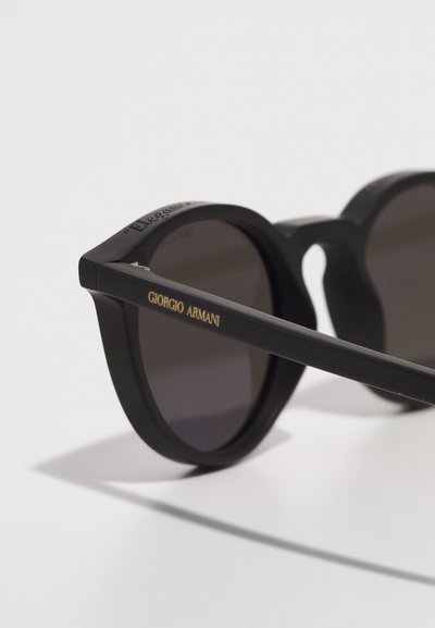 Černé kulaté sluneční brýle s matným povrchem, které mají tmavé čočky a zlaté logo "Giorgio Armani" na této straně.