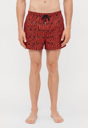 Mann in roten Badehosen mit schwarzem "DSQUARED"-Textmuster, barfuß vor einem weißen Hintergrund stehend.