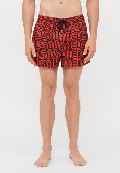 Mann in roten Badehosen mit schwarzem "DSQUARED"-Textmuster, barfuß vor einem weißen Hintergrund stehend.