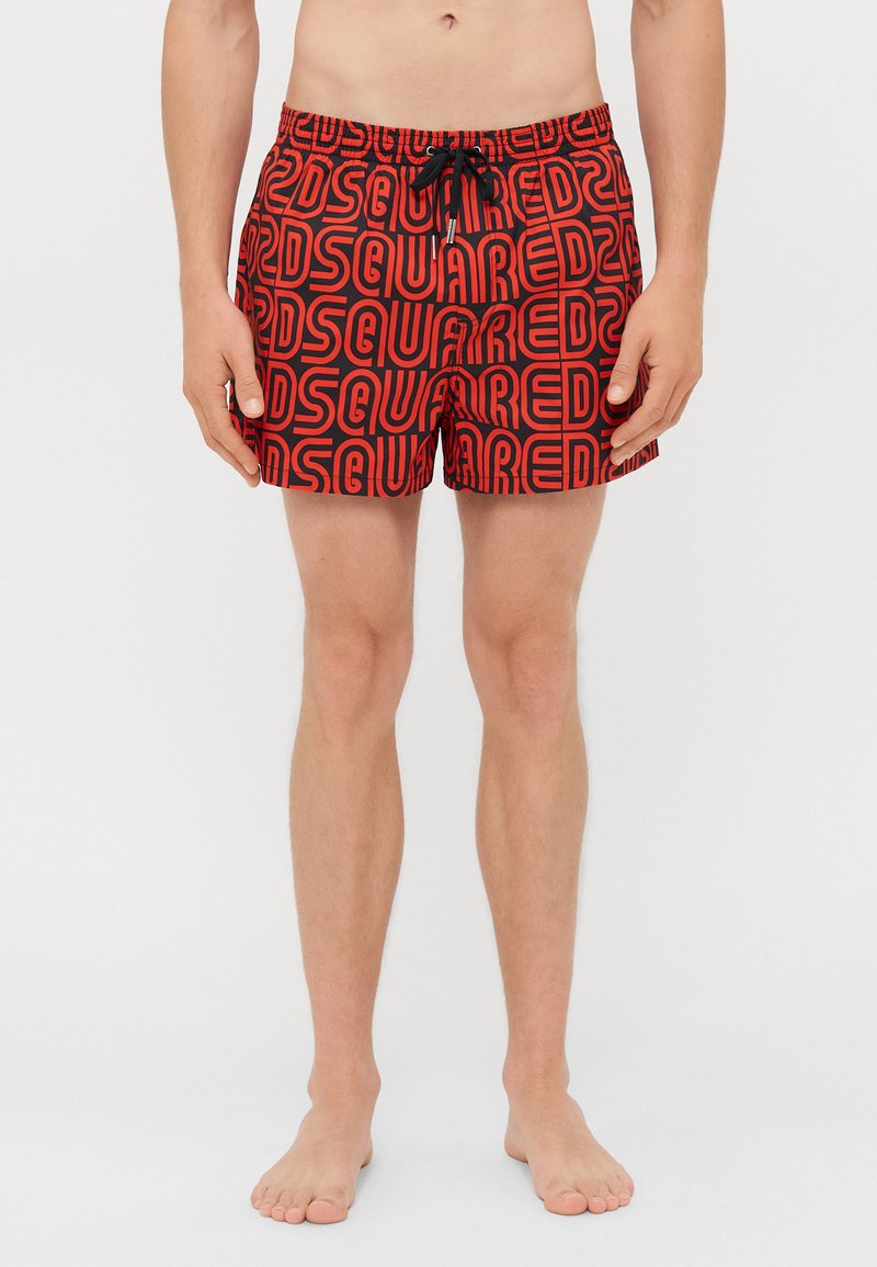 Homme portant un short de bain rouge avec un motif de texte noir "DSQUARED", debout pieds nus contre un fond blanc.