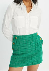 Mini gonna in tweed verde con dettagli in bottone dorato, abbinata a una camicia bianca con bottoni dotata di due tasche sul petto e un colletto casual.