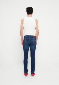 Μπλε τζιν παντελόνι με εφαρμοστή σχεδίαση, που διαθέτει καφέ δερμάτινο σημάδι στη μέση, πίσω τσέπες και φορεμένο με λευκή αμάνικη μπλούζα.