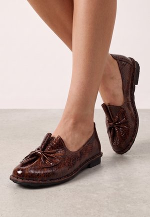 Brune blanke teksturerede læder loafers med stor forreste sløjfe, båret på bare fødder, stående på glat lysegråt gulv.